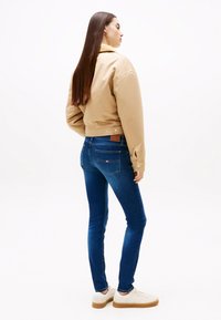 Kvinna med långt rakt hår, klädd i beige jacka, blå tajta jeans och vita sneakers, stående i en trekvart bakåtriktad pose.