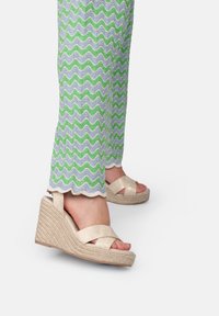 Sandalias de cuña beige con correas de tela entrelazadas, que presentan una suela de estilo alpargata texturizada. Combinadas con pantalones con un patrón y bordes en forma de concha.