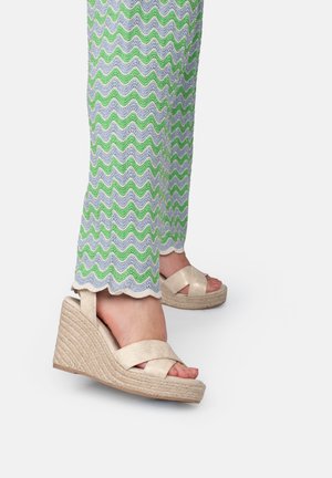 Beige Wedge-Sandalen mit kreuzgegelten Stoffriemen, mit einer strukturierten Espadrille-Sohle. Kombiniert mit gemusterten Hosen mit gewelltem Saum.