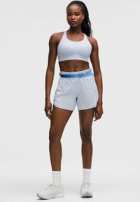 Pantalones cortos deportivos de color azul claro con cintura elástica y acentos azules, combinados con un sujetador deportivo blanco. La modelo se encuentra en un fondo neutro.