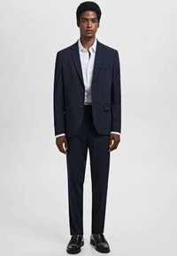 Traje navy compuesto por un blazer ajustado con un bolsillo en el pecho y pantalones de frente plano, combinado con una camisa blanca de botones y zapatos negros.