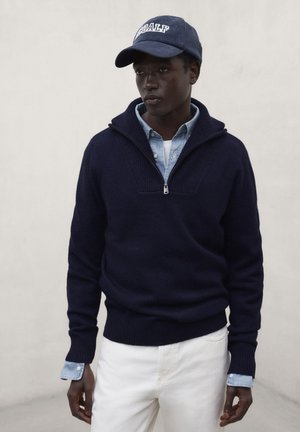 Hombre que lleva un suéter azul marino con cremallera sobre una camisa azul claro abotonada, pantalones blancos y gorra azul oscuro con letras blancas, de pie.