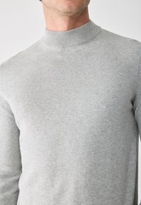 Hellgrauer Strickpullover mit hohem, geripptem Kragen und glattem, strukturiertem Finish. Nahaufnahme zeigt Schulter- und oberen Brustbereich.