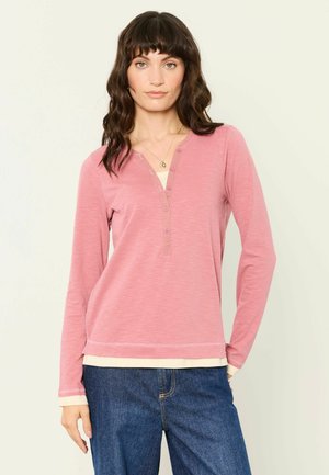 Kvinde med skulderlangt, bølget mørkt hår bærer en pink langærmet henley-shirt lagdelt over en lys cremefarvet top, kombineret med blå jeans.