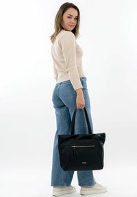 Black leather tote bag with a textured finish, featuring a front zipper pocket and sturdy fabric handles. Paired with light blue jeans and beige sweater.
