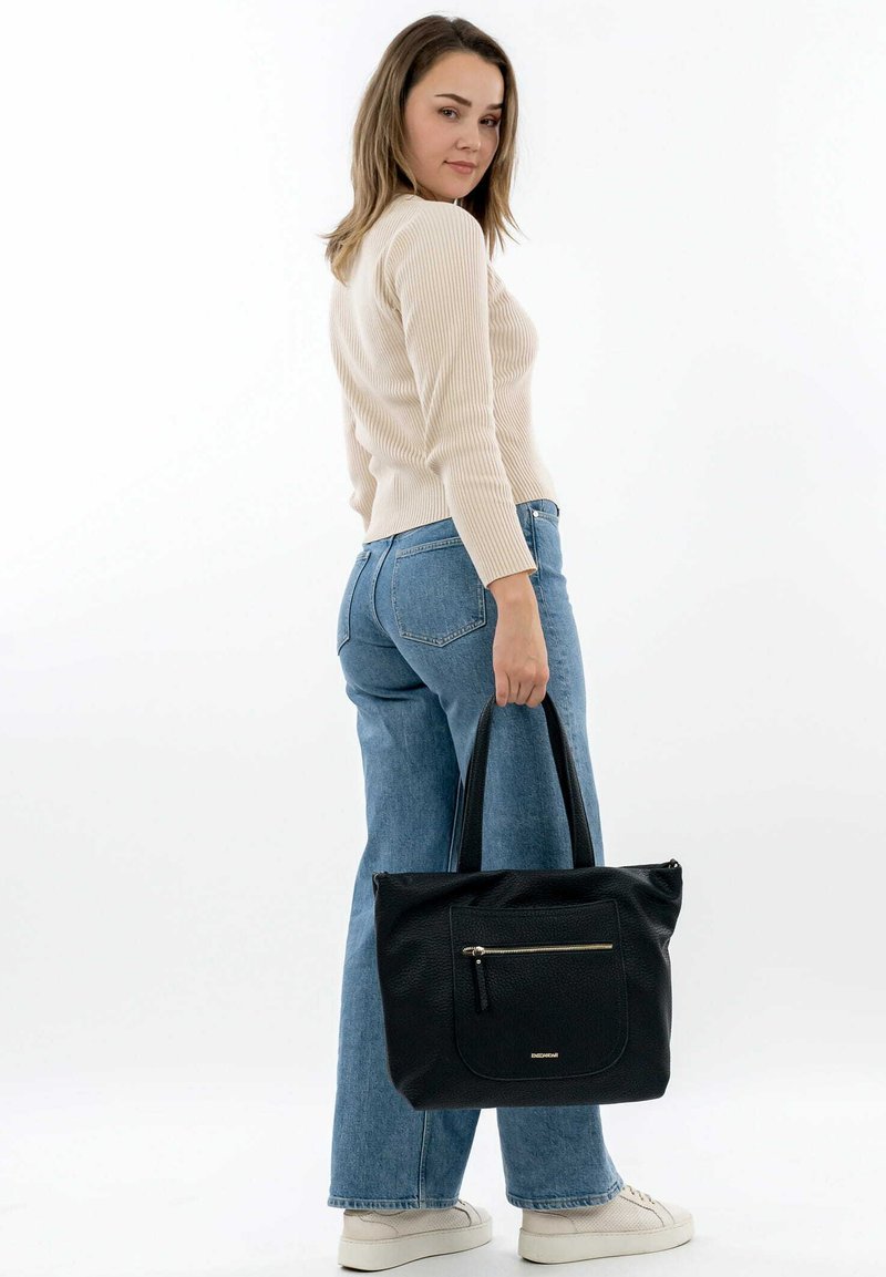 Black leather tote bag with a textured surface, featuring a front zip pocket and short handles, held by a person in light pants and sweater.