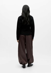 Pull noir, coupe ample. Pantalon marron, ample avec des poignets élastiques. Texture lisse, design minimaliste. Chaussures noires. Fond blanc.