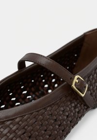 Barna szövéssel készült lapos bőr cipő Mary Jane pánttal és arany csattal, fehér háttéren fényképezve.