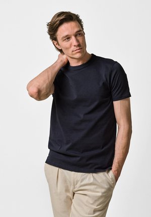 Hombre con cabello castaño que lleva una camiseta negra de manga corta y pantalones beige, con una mano apoyada en el cuello y la otra en el bolsillo.