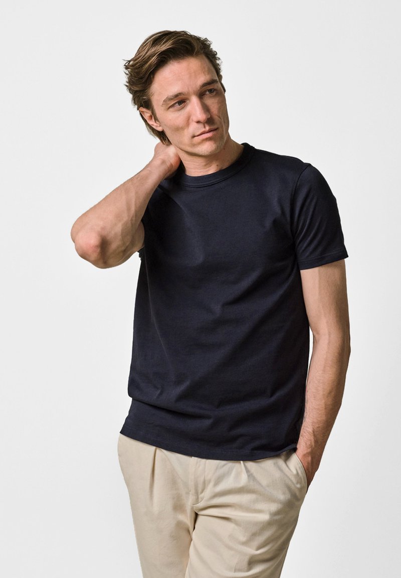 Mand med brunt hår iført en sort kortærmet t-shirt og beige bukser, hvilende den ene hånd på nakken og den anden i lommen.