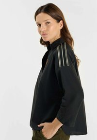 Zwarte blouse met een opstaande kraag en driekwartmouwen. Voorzien van drie verticale strepen in een lichte kleur op de schouder. Gladde textuur.