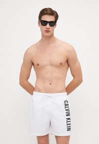 Biele plavky s čiernym logom "CALVIN KLEIN" vytlačeným vertikálne, pružný pás s šnúrkou; model nosiaci slnečné okuliare, bez trička.
