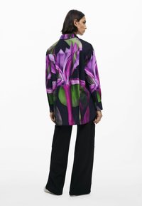 Camicia lunga con un design floreale in viola e verde su uno sfondo scuro. Abbinata a pantaloni larghi neri con tasche laterali.