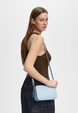 Giovane donna con lunghi capelli castani che indossa una canottiera marrone e jeans scuri, con una borsa a tracolla azzurro chiaro con una tracolla lunga.