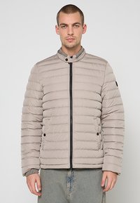 Lichtbeige donzen jas met ritssluiting aan de voorkant, horizontale stiksels, hoge kraag en drukknopaccenten. Zachte textuur en ontspannen pasvorm.