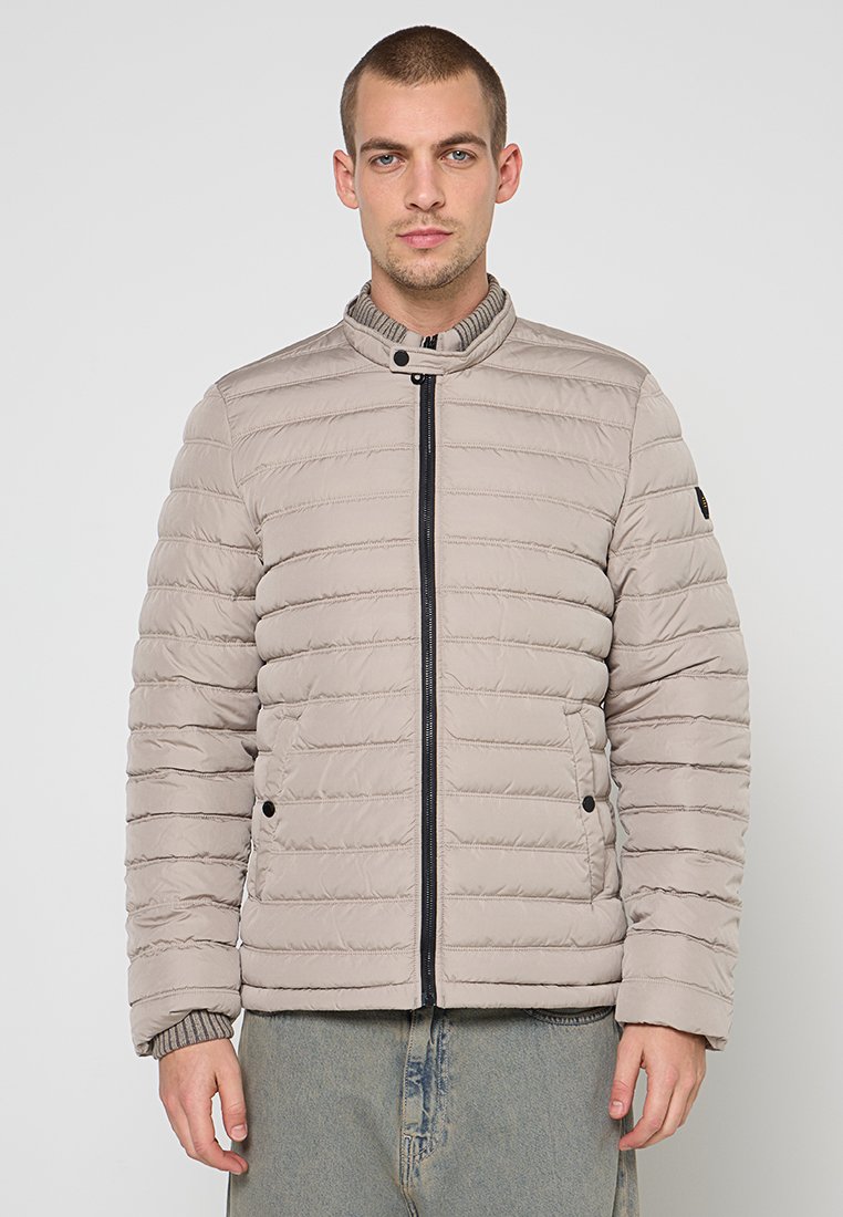 Lichtbeige donzen jas met ritssluiting aan de voorkant, horizontale stiksels, hoge kraag en drukknopaccenten. Zachte textuur en ontspannen pasvorm.