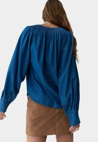Blusa in denim blu con scollatura elastica e maniche a sbuffo, abbinata a una gonna in suede marrone. Presenta bottoni ai polsini delle maniche.