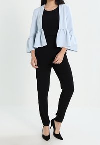 Svetle modrá peplum bunda s zvonovými rukávy přes černý overal. Modelka má na sobě černé vysoké podpatky. Hladká tkanina, střih na míru, bez vzorů.