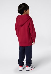 Felpa rossa con cappuccio in cotone, abbinata a pantaloni della tuta navy. La calzatura presenta una base bianca con accenti rossi e neri.