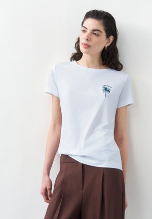 Sötét göndör hajú nő fehér rövid ujjú inget visel, amelyen apró zöld pálmafák láthatók, és barna, magas derekú, bő szárú nadrágot, miközben a falnak dől.