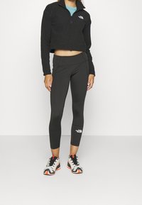 Chaqueta de felpa negra recortada con media cremallera, llevada sobre una blusa azul verdosa; combinada con leggings negros con un logo y costuras planas; zapatillas blancas y naranjas.