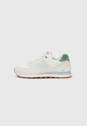 Beige and white New Balance sneaker with light green "N" logo, green heel tab, white midsole, and gum rubber outsole on gray background.