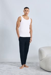 Μυώδης άνδρας που στέκεται ξυπόλυτος πάνω σε χαλί, φοράει λευκό αμάνικο μπλουζάκι και μαύρο παντελόνι, δίπλα σε καμπύλο, υφασμένο λευκό καναπέ.