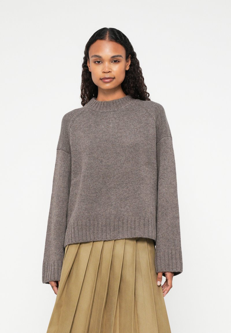Brown knitted sweater with a crew neck, ribbed hem, and dropped shoulders. Soft texture, paired with a pleated, beige skirt.