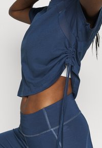 Nära bild av person i marinblå träningsoutfit med rynkade sidodetaljer på toppen, som visar midjan och en upplyft arm.