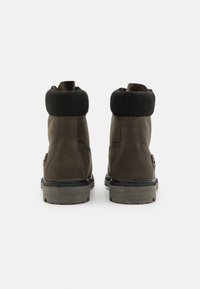 Botine din piele maro până la gleznă cu mansetă din velur negru, suprafață texturată, cusături consolidate, talpă din cauciuc gri rezistentă și detaliu de branding subtil.
