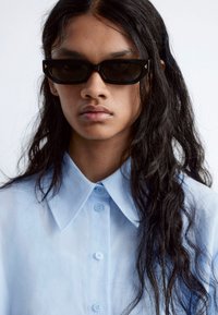 Black sunglasses with a cat-eye shape, dark lenses, and subtle accents, paired with a light blue button-down shirt featuring a pointed collar.