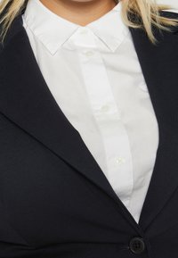 Blazer noir avec une texture lisse, présentant une chemise blanche à boutons en dessous, montrant un col classique et deux boutons visibles.