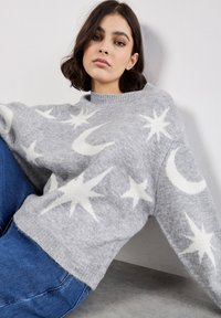 Grauer Wollpullover mit weißem Stern- und Mondmuster, ausgestattet mit einem gerippten Rundhalsausschnitt und lockerer Passform, kombiniert mit blauen Jeans.