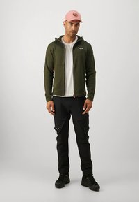 Žalia striukė su užtrauktuku dėvima ant baltos marškinėlių, derinama su juodomis krovinių kelnėmis ir rožine kepure. Svarbūs bruožai yra kišenės ir tekstūruota medžiaga.