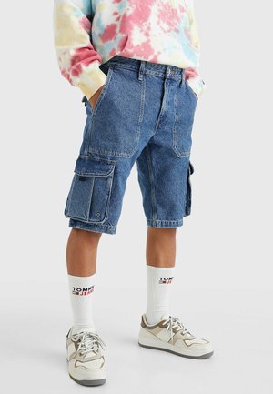Person iført blå denim cargoshorts, hvide Tommy Jeans crew sokker, beige og hvide sneakers og en flerfarvet tie-dye sweatshirt.