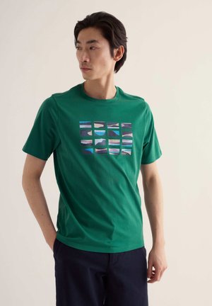 Joven con camiseta verde con una cuadrícula de diseños abstractos de montañas y mar, de pie con una mano en el bolsillo, mirando de lado.