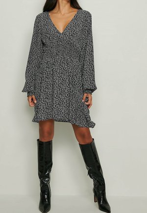 Woman wearing a black and white floral V-neck dress with long sleeves paired with knee-high black patent leather boots.