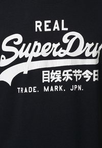 Černé bavlněné tričko s velkým bílým textem, který zní "REAL SuperDry", a dalším bílým textem v menším písmu nad a pod ním.