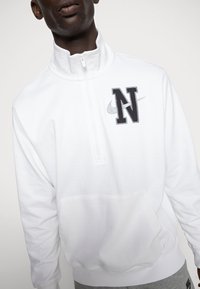 Sudadera blanca con cremallera y cuello alto, que presenta un bordado negro de la 'N' y un logo de Nike en gris. Incluye un bolsillo frontal y dobladillo acanalado.