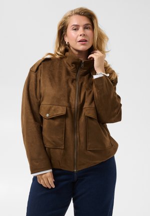 Femme blonde portant une veste en daim marron avec des poches avant et un pantalon bleu marine, ajustant le col de la veste d'une main.