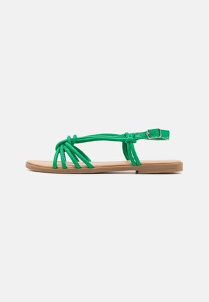 Sandalias planas verdes con tiras anudadas, con suela marrón y hebilla ajustable en el tobillo, que presentan un diseño minimalista.