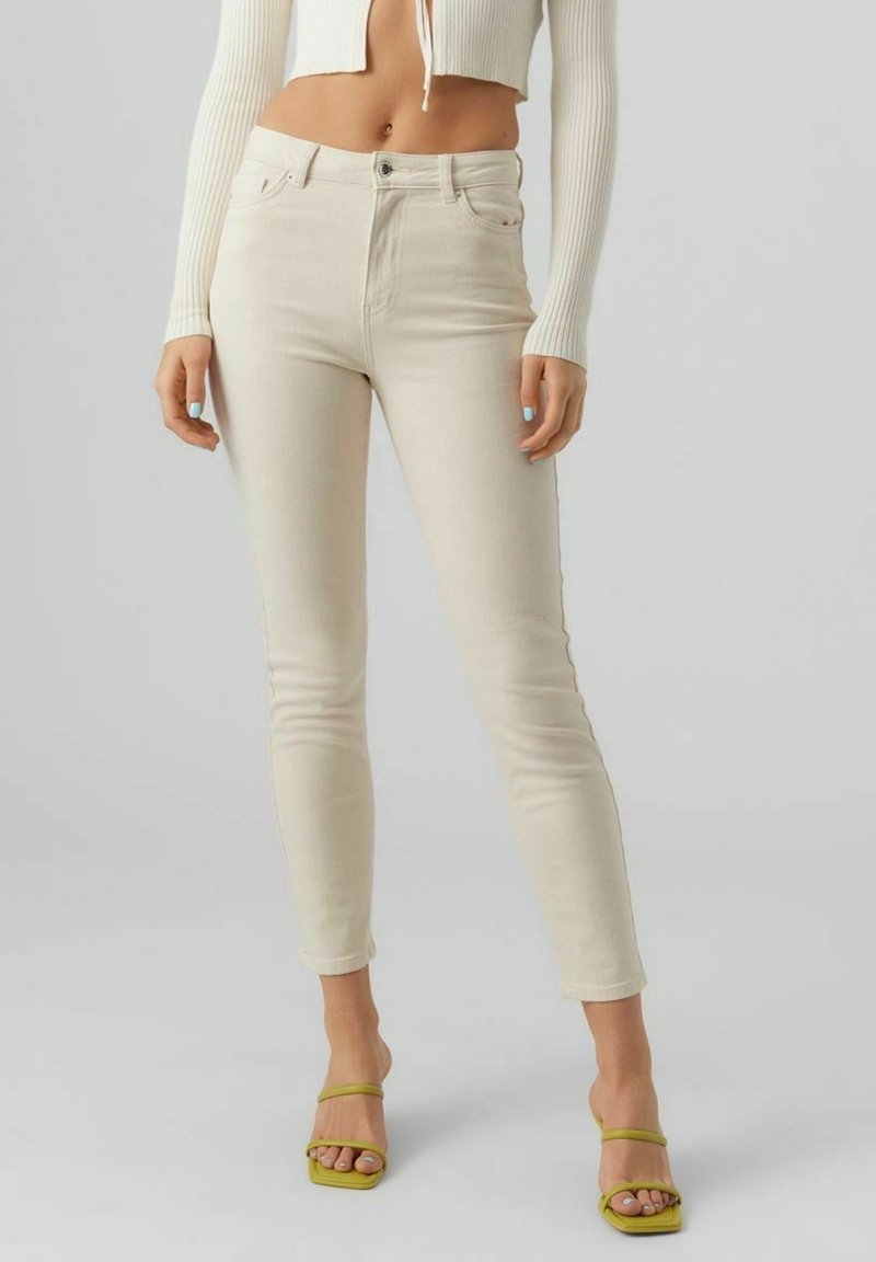 Femme portant un jean skinny taille haute couleur écru, un top court côtelé crème et des sandales à talons ouvertes vertes, debout devant un fond uni.
