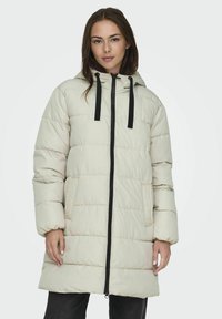 Doudoune beige clair avec capuche, fermeture éclair noire et cordons de serrage. Présente un design matelassé et des poches latérales.