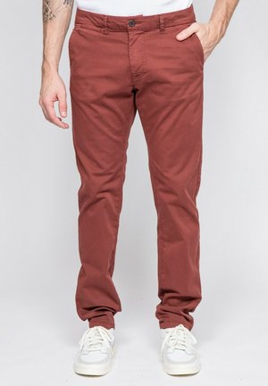 Homme portant un pantalon chino couleur rouille, des baskets blanches et une chemise blanche, avec une main dans la poche et un tatouage visible sur le bras gauche.