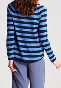 Sininen ja laivastonsininen raidallinen neulepaita, jossa pitkät hihat, pehmeä rakenne, pyöreä pääntie ja kaareva helma, yhdistetty vaaleanharmaisiin housuihin.