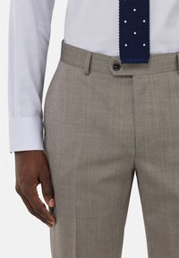 Pantalones de rayas beige con un frente plano, que cuentan con un cierre de botón. Combinados con una camisa de cuadros azul claro y una corbata de lunares en azul marino.