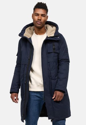 Parka larga azul marino con capucha forrada de pelaje sintético, bolsillos con cremallera y cierre frontal. Llevada sobre un sudadera color crema y jeans de mezclilla.
