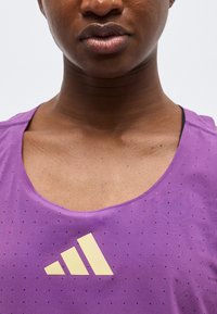 Κοντινό πλάνο του κάτω μέρους του προσώπου και του λαιμού ενός ατόμου που φοράει μοβ τοπ χωρίς μανίκια με κίτρινο λογότυπο Adidas.