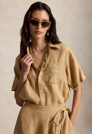 Γυναίκα που φορεί καφέ κοντομάνικο πουκάμισο με κουμπιά και φούστα που ταιριάζει με δέσιμο στη μέση, πράσινο κολιέ με χάντρες, σκουλαρίκι με κοχύλι και γυαλιά ηλίου με σχέδιο καβούρι.