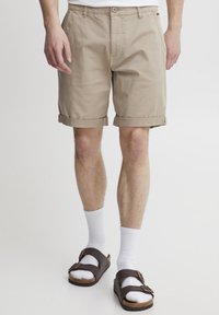 Beige Baumwollshorts mit umgeschlagenem Saum, ausgestattet mit Fronttaschen, getragen mit weißen Socken und braunen Sandalen mit zwei Schnallen.